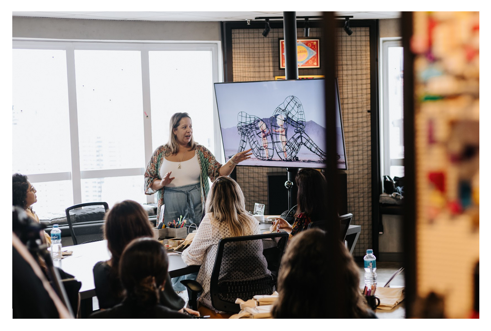 Alessandra Miliatti apontando para uma televisão onde está sendo exibido um dos slides da aula, enquanto algumas mulheres prestam atenção sentadas em volta de uma mesa.