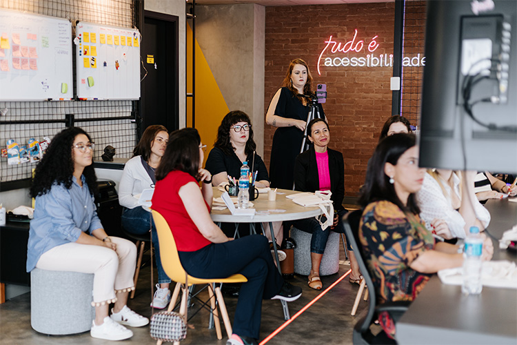 A câmera está posicionada atrás da televisão onde está sendo exibidos os slides da aula e ao fundo todas as mulheres aparecem sentadas em volta de uma mesa.