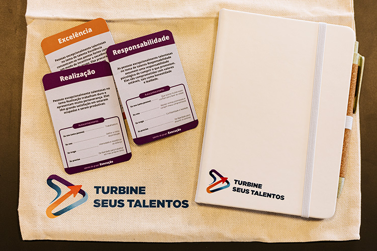 Kit de boas-vindas que todas as participantes receberam, contendo um bloco de anotações, uma caneta, uma sacola retornável e um baralho de talentos.